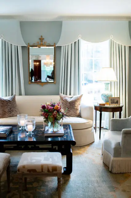 Living room with a cream sofa, tiger print pillows, a black lacquered coffee table with a floral arrangement and candles, and light blue drapes.