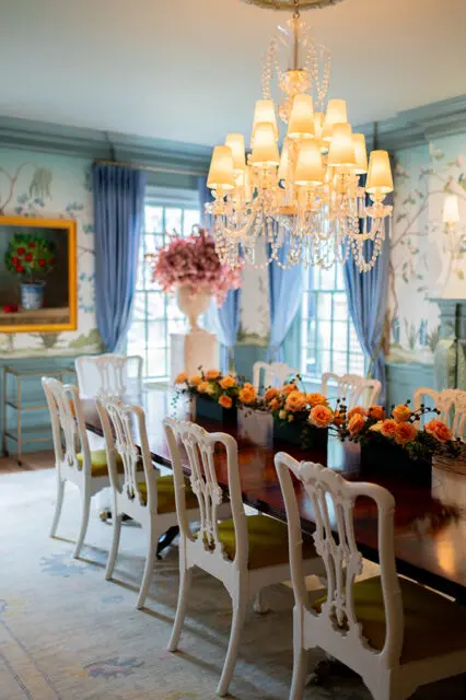 Formal dining room with a dark wood table set with orange floral arrangements, white chairs with green upholstery, a crystal chandelier, and chinoiserie wallpaper.