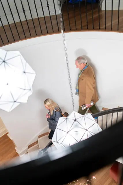 A woman in a grey jacket and a man in a tan coat walk up a curved staircase with white walls, black railings, and geometric pendant lights.