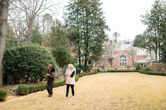 Two women walk across a large lawn towards a red brick Georgian-style mansion with a manicured garden and trees.