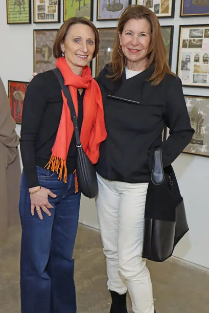 Katie Johnson and Catherine Cage stand in front of a wall of framed art. Johnson wears a black sweater, orange scarf, and blue jeans. Cage wears a black jacket, white pants, and black boots.
