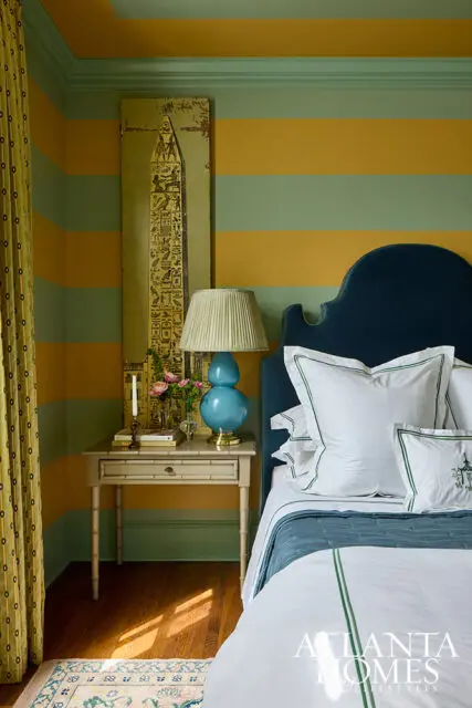 Guest bedroom with horizontal stripes of mustard yellow and sage green on walls and ceiling. Features a navy velvet headboard, white bedding with green trim, a bamboo-style nightstand with a blue lamp, and Egyptian-themed artwork.