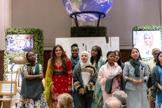 A group of girls stand on a stage in front of screens displaying images and a large globe. One girl wears a red and gold dress, another a hijab and striped sweater, and others wear colorful clothing.