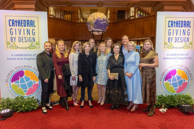 A group of eleven people stand in front of banners that read "Cathedral Giving by Design" and "Global Village Project." A large globe is visible behind them.