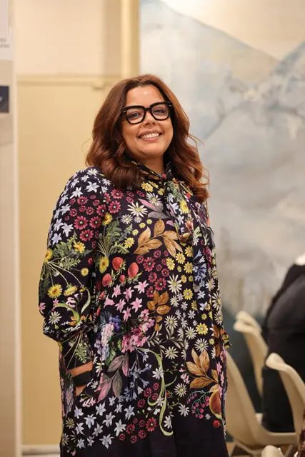 Woman with auburn hair and glasses smiles, wearing a dark dress with a colorful floral print.