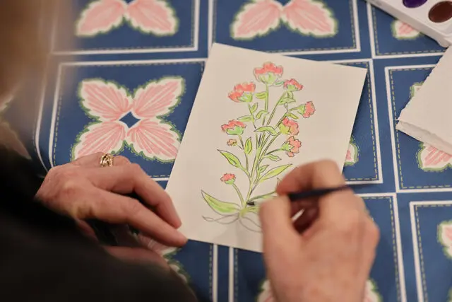 Close-up of hands painting a floral illustration with watercolors on a blue patterned tablecloth.