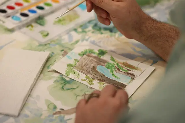 Close-up of a person's hands painting a watercolor landscape featuring a bridge and water, with a palette of watercolors and a brush nearby.