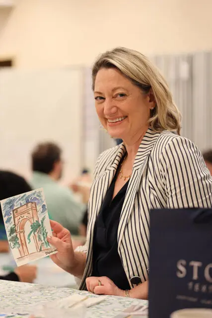 Woman in a striped blazer holds a watercolor painting of an archway and smiles at the camera.
