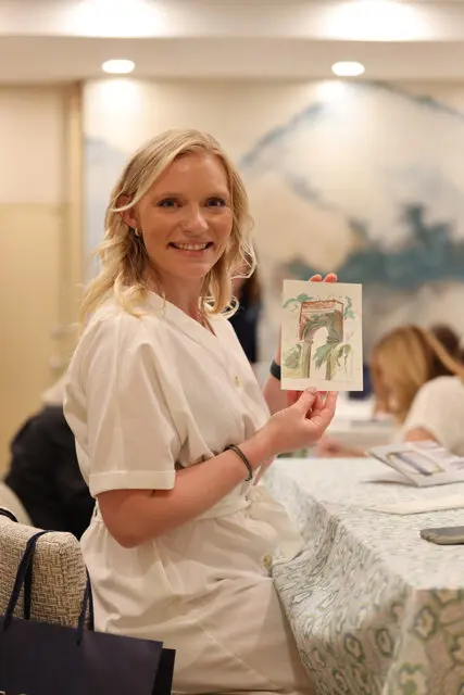 Woman with blonde wavy hair wearing a white dress, holding up a small watercolor painting of an arched entryway with greenery.