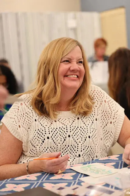 Woman with blonde hair smiling and holding a paintbrush, wearing a cream-colored crocheted top.