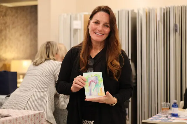 Woman with auburn hair smiling and holding a small watercolor painting of an arched structure with greenery. She wears a black cardigan over a black top and patterned pants.
