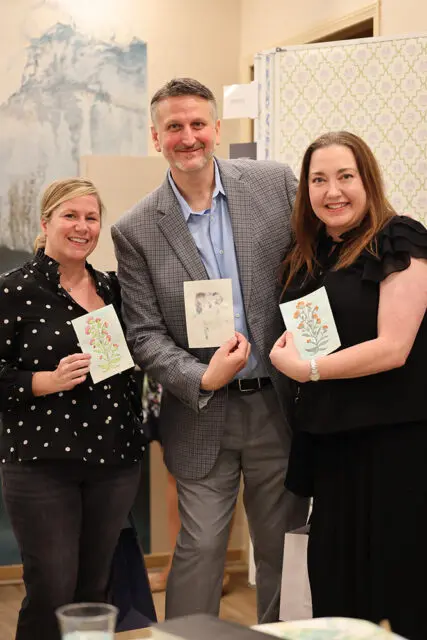 Three people smiling and holding up small artworks. The woman on the left holds a floral illustration. The man in the middle holds a sketch of two figures. The woman on the right holds an illustration of an orange-flowered branch.