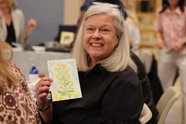 Woman with gray hair smiling and holding a watercolor painting of yellow flowers. She is wearing a black shirt and glasses.