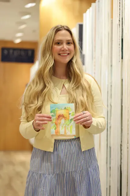 Young woman with long blonde hair wearing a yellow cardigan and blue and white striped skirt, holding a watercolor painting of an arched structure with greenery.