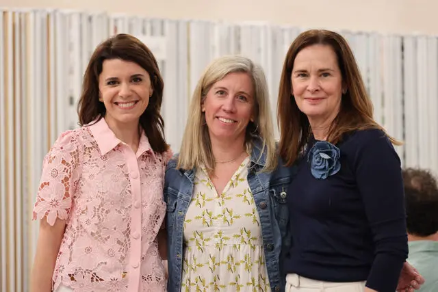 Three women smiling at the camera. The woman on the left wears a pink lace shirt. The woman in the middle wears a white dress with a green leaf pattern and a denim jacket. The woman on the right wears a navy sweater with a blue fabric flower on the lapel.