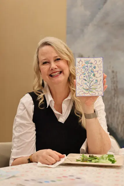 Woman with blonde hair smiling and holding up a small painting of a floral design. She is wearing a white shirt and black vest.