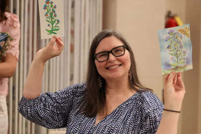 Woman with glasses smiling and holding up two watercolor paintings of flowers.