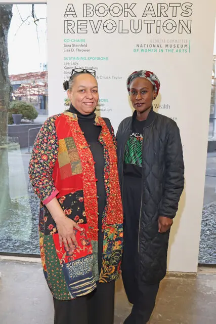 Two women stand in front of a banner that reads "A BOOK ARTS REVOLUTION" and lists sponsors and "GEORGIA COMMITTEE NATIONAL MUSEUM OF WOMEN IN THE ARTS." The woman on the left wears a colorful patchwork jacket.