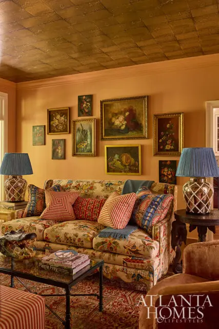 Living room with peach walls, a patterned sofa with colorful pillows, a glass coffee table with books and an alligator sculpture, and a gold tiled ceiling.