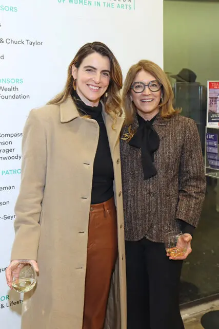 Two women stand together, each holding a glass of white wine. The woman on the left wears a tan coat and brown pants. The woman on the right wears a textured brown jacket and black pants.