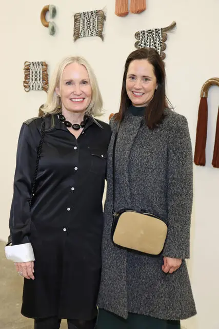 Lisa Cannon and Robin Aiken posing in front of woven wall art. Lisa wears a black satin shirt and Robin wears a grey textured coat.