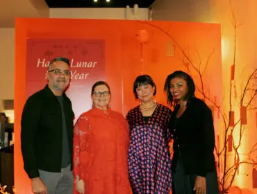 Four people stand in front of an orange backdrop with Lunar New Year decorations. A man in a dark shirt and grey pants smiles. A woman in a bright orange lace dress smiles. A woman in a patterned dress and gold necklace smiles. A woman in a black cardigan smiles.