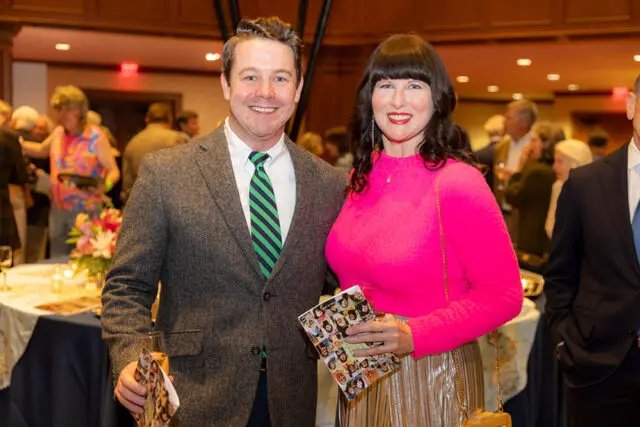 Man in a grey tweed jacket and green striped tie smiles next to a woman in a bright pink sweater and gold pleated skirt.