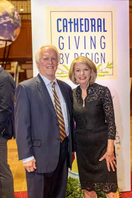 Man in a navy suit and striped tie stands next to a woman in a black lace dress in front of a sign that reads "CATHEDRAL GIVING BY DESIGN"