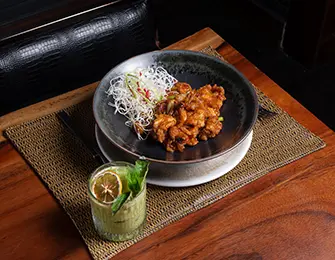 A dark-rimmed bowl of glazed chicken pieces topped with shredded white vegetables sits on a woven placemat next to a glass of green cocktail garnished with a lemon slice and mint leaves.