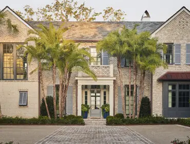 Brick home with gray shutters and a copper roof bay window, featuring a covered entryway with palm trees and manicured landscaping.
