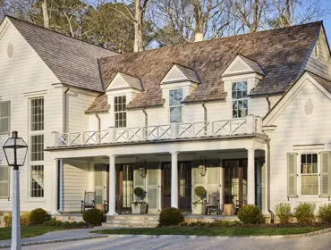 White clapboard house with a cedar shake roof, dormer windows, and a wraparound porch with white railings and columns.