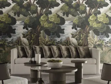 Living room with a cream sofa adorned with velvet pillows, set against a scenic wallpaper featuring trees, water, and a heron. Round wooden coffee tables and textured chairs complete the space.