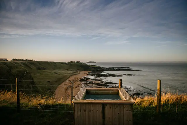 Wooden hot tub overlooking a rocky coastline and the ocean at sunset