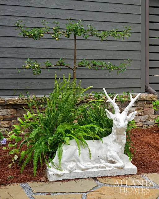 White deer statue among ferns and a small apple tree against a gray wood-paneled wall.