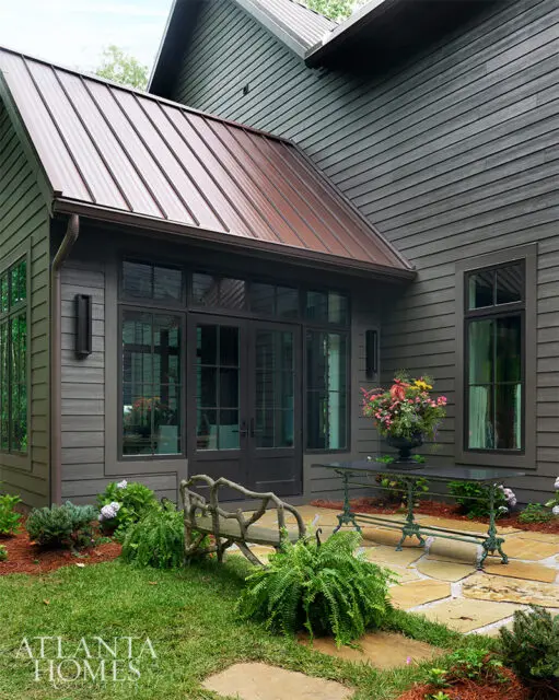 Dark gray siding, copper roof, French doors, stone patio, bench, and table with flowers.