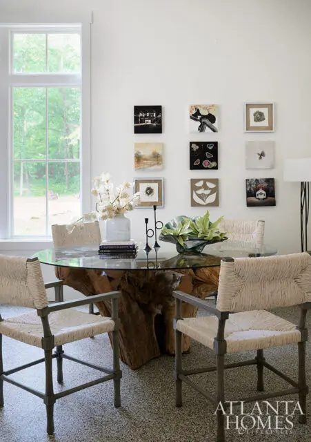 Dining room with glass-top table on a wood base, woven chairs, and nine framed artworks on wall.