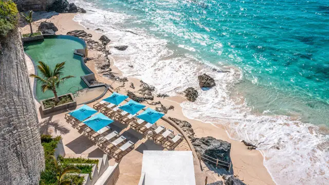 Oceanfront infinity pool with lounge chairs and blue umbrellas on a sandy beach.