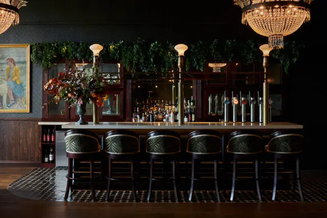 Bar with upholstered stools, liquor bottles, and beer taps. Dark wood paneling and a painting on