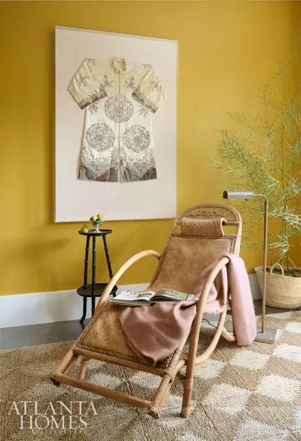 Rattan lounge chair with pink throw, book, and glasses. Yellow wall with framed robe.