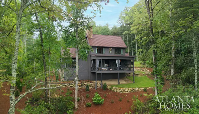 Dark gray house with brown metal roof and stone chimney, surrounded by trees.