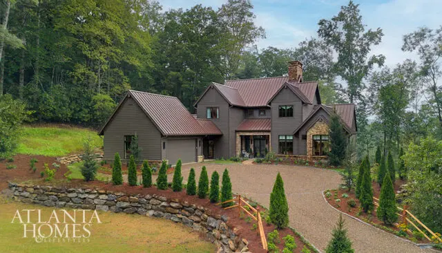 Brown house with metal roof, stone accents, and gravel driveway surrounded by trees.