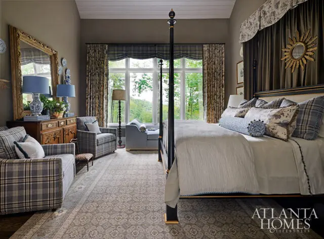 Bedroom with four-poster bed, plaid armchairs, and large window overlooking trees.