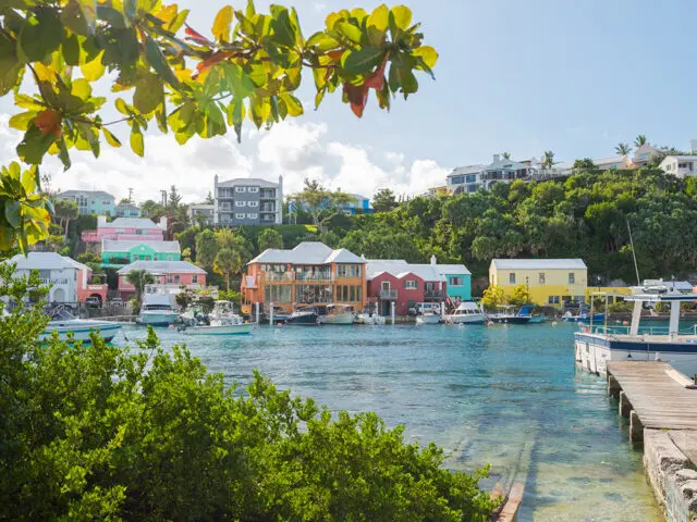 Colorful houses line a harbor with boats docked and lush green hills in the background.