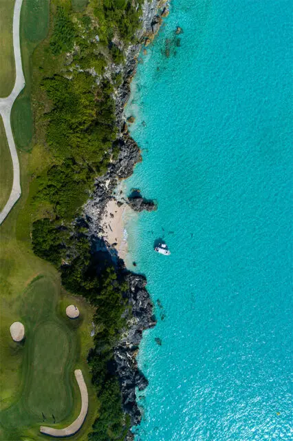 Aerial view of a golf course bordering a rocky coastline and turquoise ocean with a boat.