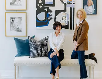 Two women in a hallway with artwork on the walls and a bench with decorative pillows.