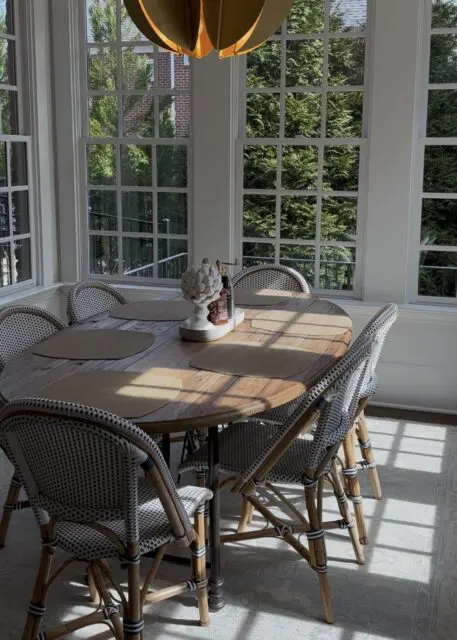Dining room with oval wood table, wicker chairs, and large windows overlooking trees.