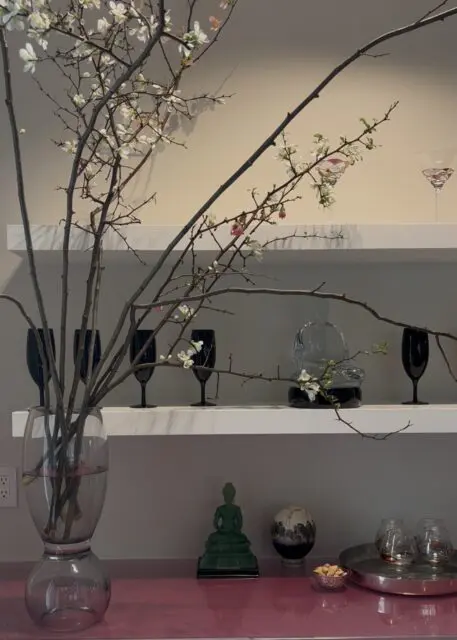 White blossoms on branches in vase, black glasses, glass Buddha, and globe on shelves.