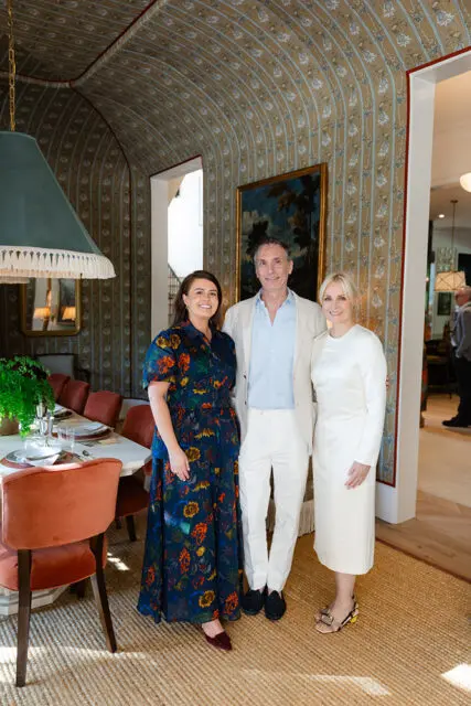Three people stand in a dining room with arched, patterned wallpaper and a long table set for