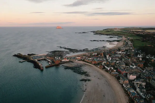 Coastal town with harbor and beach at sunset, with a small island offshore.
