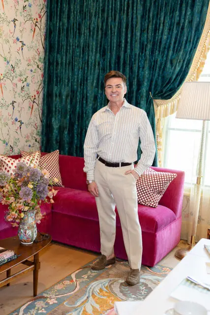 Man in striped shirt and tan pants standing by a fuchsia sofa with patterned pillows.